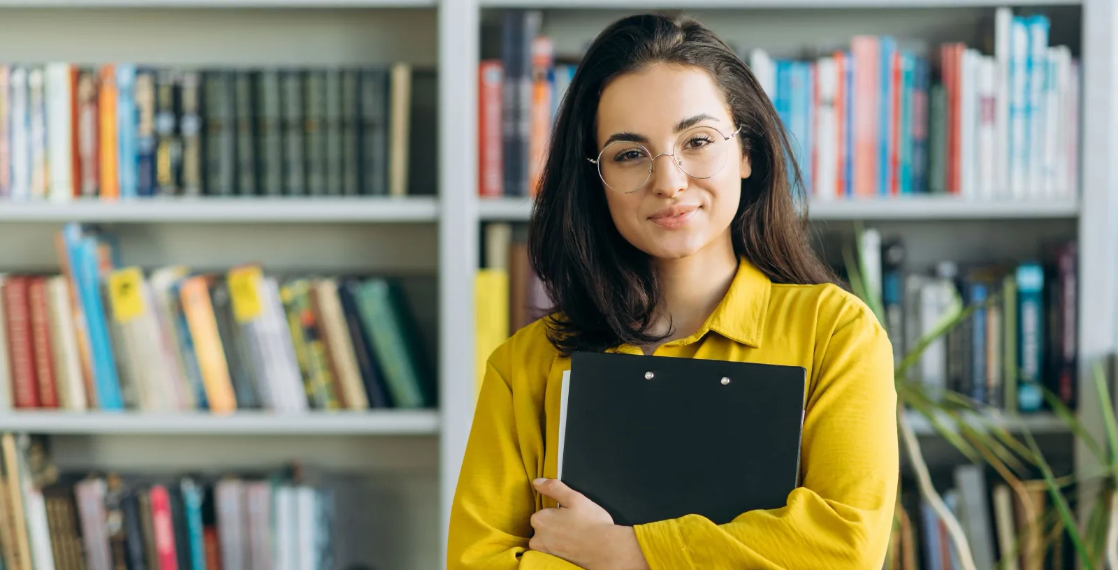A university lecturer holding a clipboard, standing in front of library shelves.